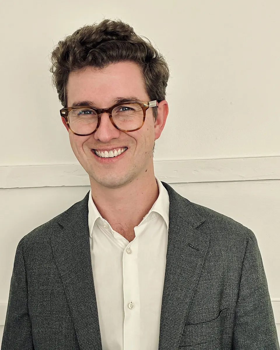 Tristan Rolls in tortoiseshell glasses and a grey blazer, smiling against a cream wall